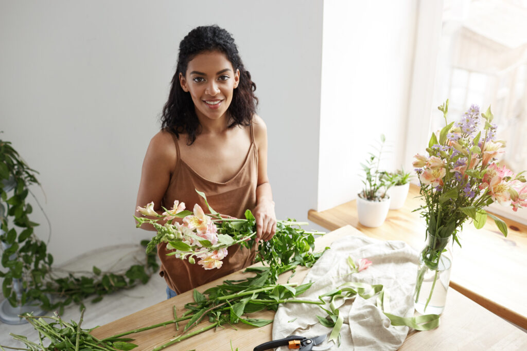 young beautiful african girl florist taking care of flowers at workplace over white wall.